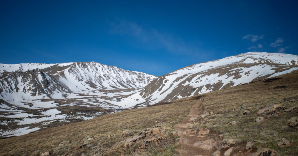 Dirt trail crossing alpine tundra on Mount Elbert with snow-covered slopes beyond